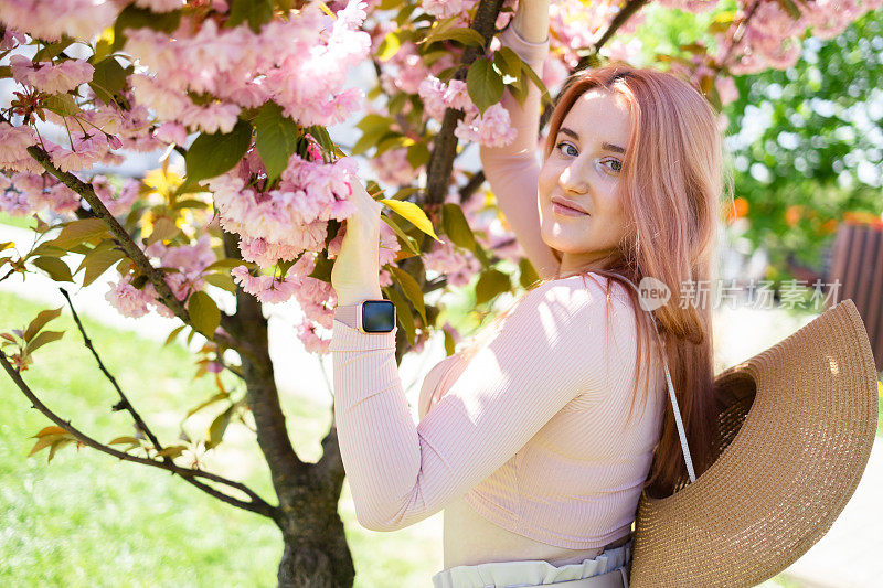 美丽的女孩在樱花树旁。女人穿着裙子和时髦的外套粉红色的花朵盛开。在开花。春天的时间。放松和幸福的概念。