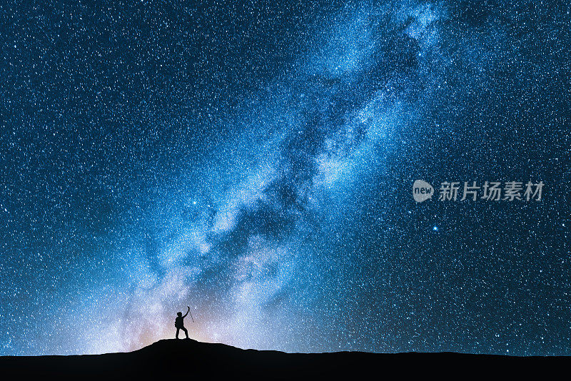男人的剪影与徒步杆对抗惊人的银河在夜间。空间背景。山水有山人，银河明亮，天空有星星。美丽的星系。旅行。蓝色星空