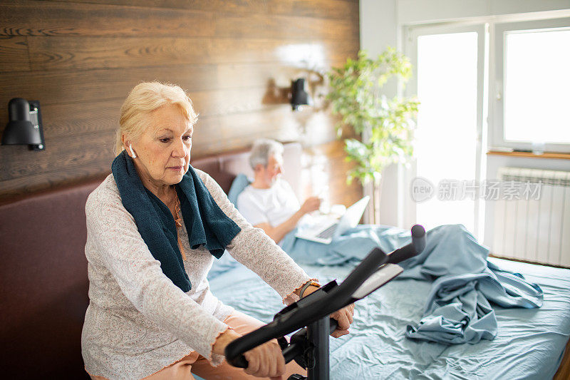 一位老年妇女正在室内自行车上锻炼，而她的丈夫正在她身后的床上使用笔记本电脑