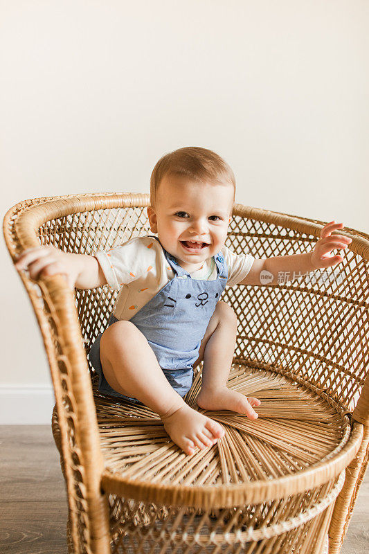 一个15个月大的古巴裔美国男婴，浅棕色短发，棕色眼睛和12只脚趾，穿着胡萝卜连体衣和蓝色兔子连体裤，还有蓬松的兔子尾巴，光着脚坐在复古藤椅上