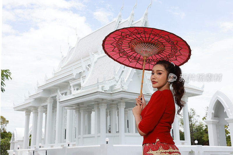 美丽的缅甸女孩在脸颊上涂抹田中粉，撑着红色的伞在寺庙里散步。一名身着红色民族服装的缅甸妇女撑着一把红色雨伞走在泰国一座白色的佛教寺庙里
