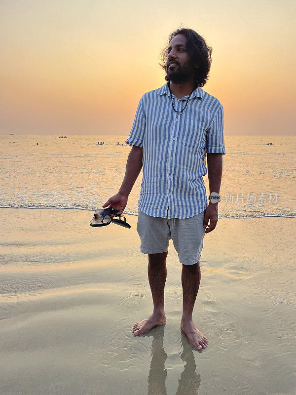 印度果阿邦帕洛莱姆，一个穿着短裤和条纹短袖衬衫的印度男子在沙滩上拿着凉鞋的特写照片，黄昏时分，太阳落山，度假游客赤脚站在海边，这是前景的焦点