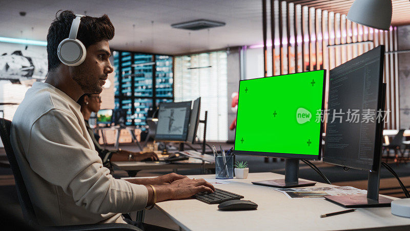富有创造力的印度人的肖像坐在桌子上使用桌面电脑与模拟绿屏Chromakey。在游戏设计初创公司工作的男性关卡设计师，创造沉浸式游戏玩法