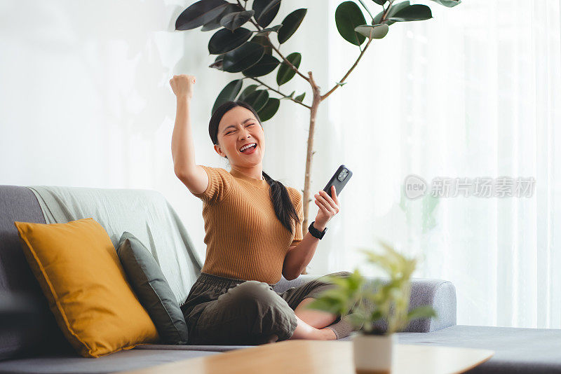 亚洲女人坐在家里的客厅里，用智能手机开心兴奋地做了一个获胜的手势。