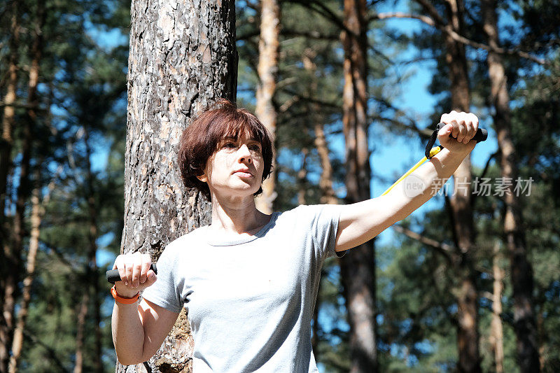 成熟的女人在松林里与阻力带一起锻炼