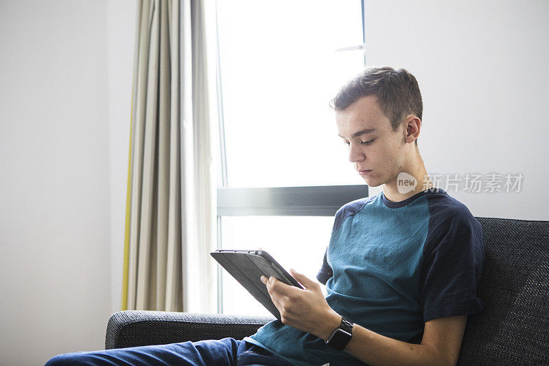 一名年轻的男学生正坐在灰色的沙发上使用平板电脑。他穿着一件蓝绿色的t恤和牛仔牛仔裤。
