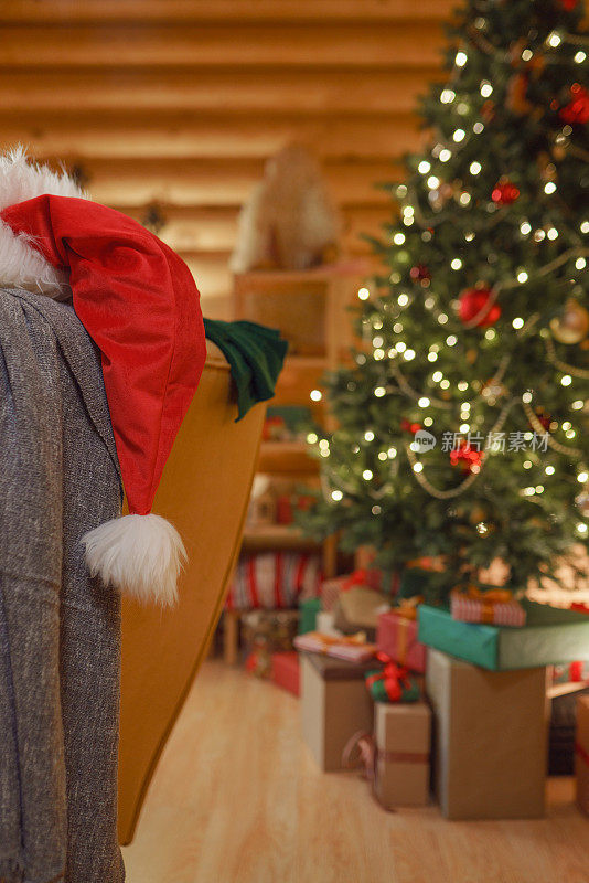 一幅圣诞画，前景的焦点是装饰模糊的圣诞树和扶手椅上的圣诞老人的帽子