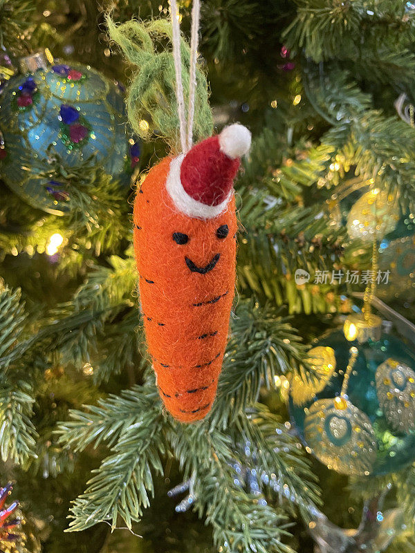 形象的毛毡胡萝卜装饰戴圣诞帽挂在人工圣诞树树枝，云杉针，模糊的白色神仙灯的背景