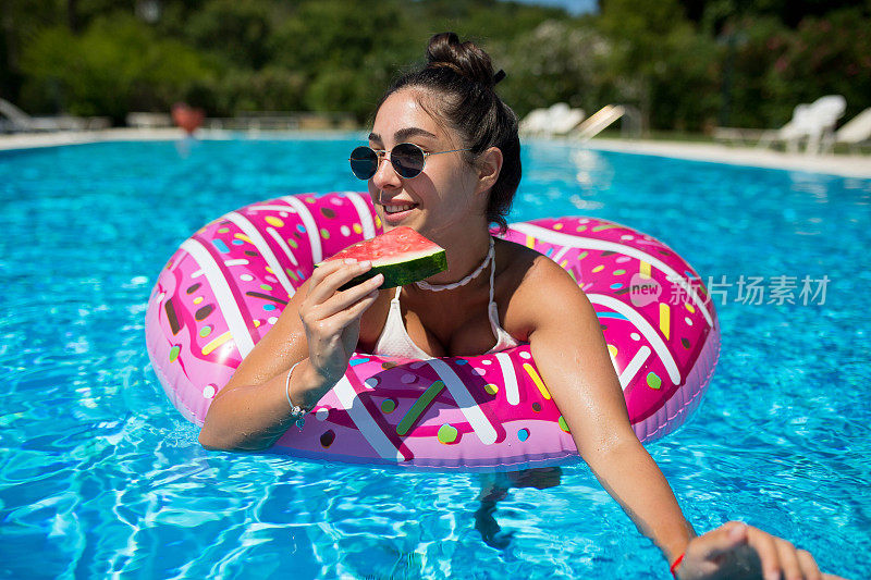 一个美丽的微笑的女孩在游泳池吃西瓜，享受一个炎热的夏天