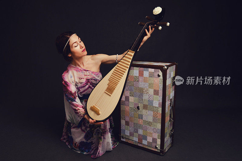 一个快乐的女音乐家穿着亚洲服装在工作室的黑色背景。黑色的背景上是一个微笑的女人，手里拿着来自亚洲的弦乐器