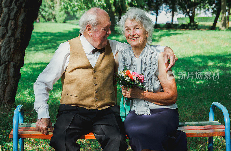 一对老夫妇坐在绿色公园的长凳上。祖母和祖父在他们的金婚纪念日上。五十年在一起。老人的爱情故事。老人欢笑拥抱