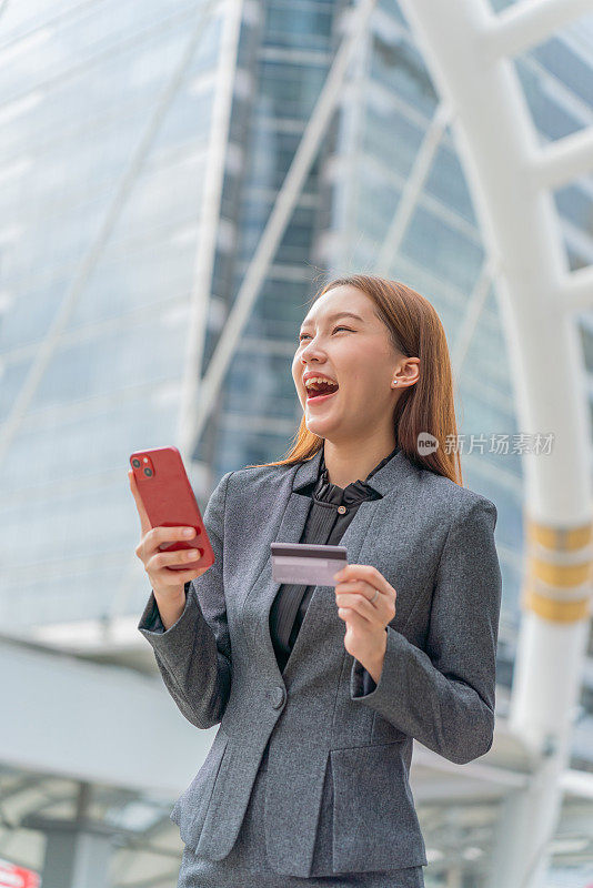 一名年轻的亚洲女商人穿着灰色的办公套装，在商业区用她的智能手机使用信用卡进行在线交易，她微笑着自信