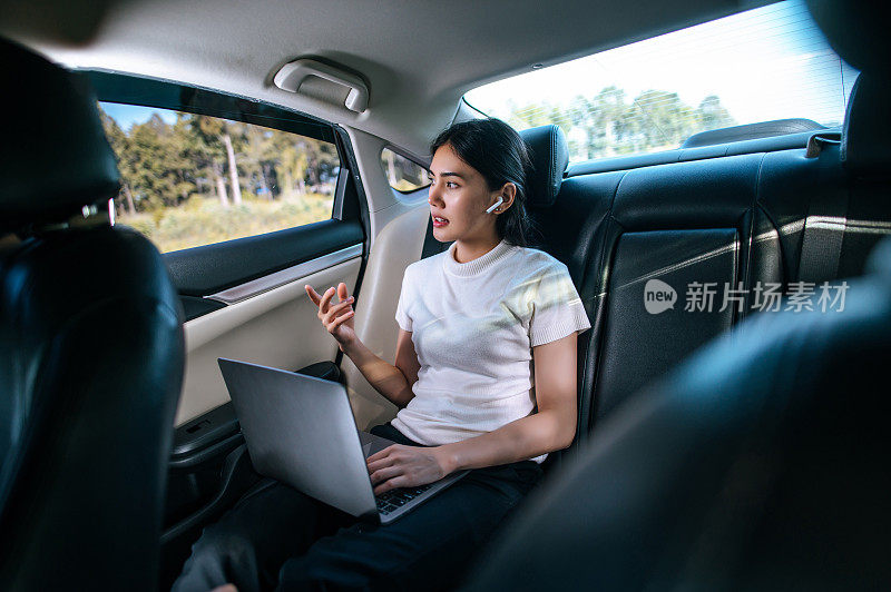 年轻的女商人在车里工作时使用笔记本电脑和无线耳机