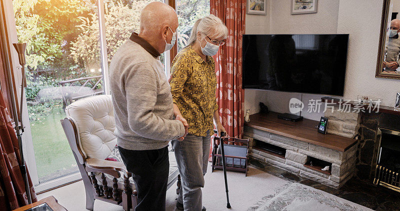 老人，夫妇或帮助和拐杖的病人，支持或协助在家里客厅的沙发上。老年人、戴口罩或患有残疾或新冠肺炎的女性与男性一起在退休休息室和康复室