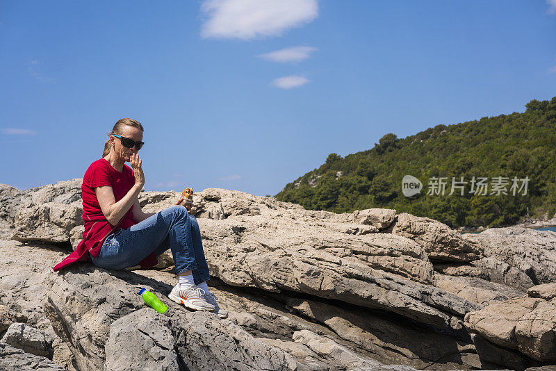 克罗地亚Mljet岛，一位资深女性坐在岩石上吃三明治