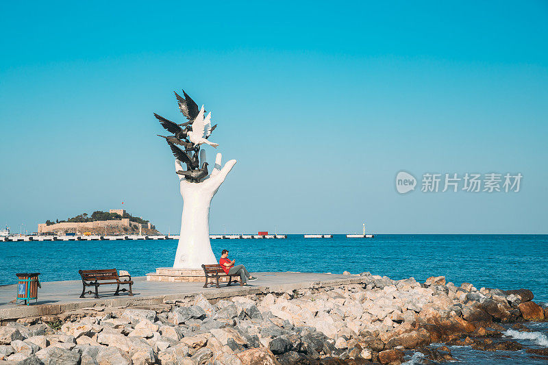 Kusadasi，艾丁省，土耳其。阳光明媚的夏日，一名男子坐在海滨和平手纪念碑旁的长凳上。土耳其爱琴海海岸的手雕塑