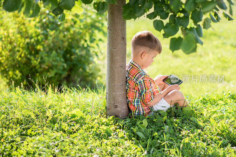一个美丽的孩子坐在草地上看手机在夏天日落。手里拿着手机的可爱男孩