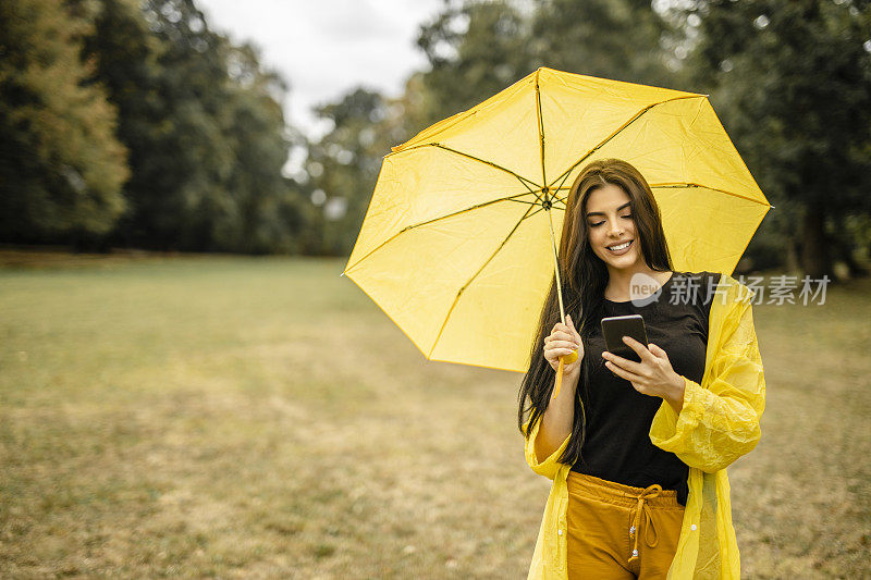 美丽的女人穿着黄色的雨衣，拿着黄色的雨伞，使用她的手机，享受在雨天