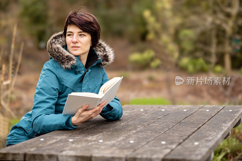 一个穿着蓝色雨衣的年轻女子坐在户外徒步旅行者野餐桌上的长椅上看书