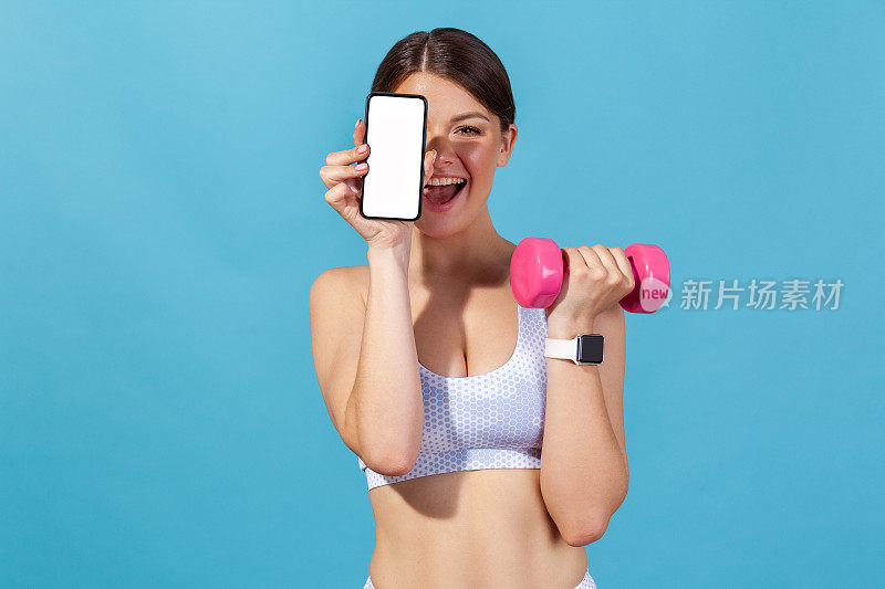 快乐的运动女性在白色运动上衣拿着空屏幕智能手机和粉色哑铃，做健身和普拉提锻炼看教程在手机上