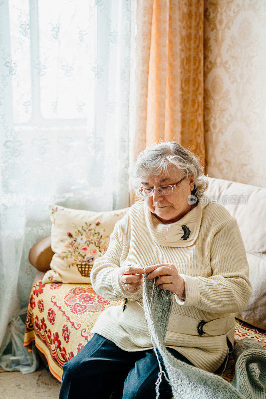 白发苍苍的老妇人正在织围巾。退休的老妇人。年迈的妻子穿着舒适的家庭毛衣。的养老金。阖家幸福长寿