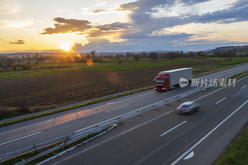 红色的卡车和轿车在公路上高速行驶，穿过乡村风景。高速公路高速行驶。高速公路上超速行驶的景象。背景是美丽的日落。
