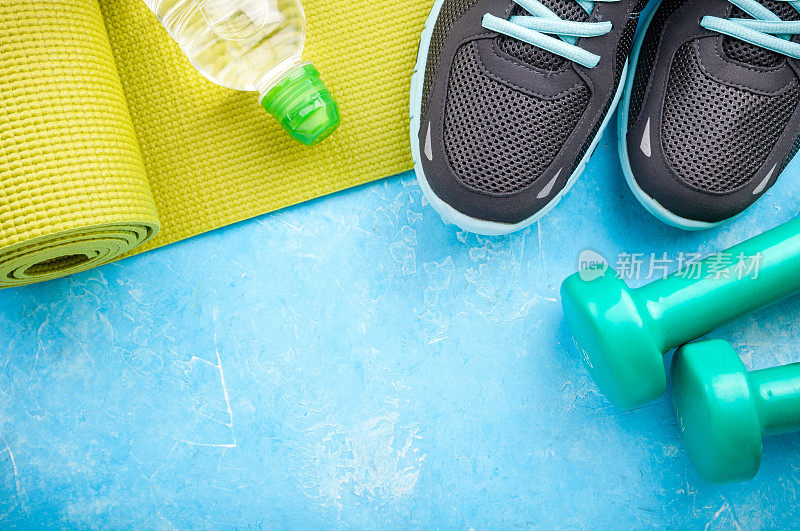 蓝色背景上的瑜伽垫、运动鞋、哑铃和一瓶水。健康生活方式、运动和饮食的概念。运动设备