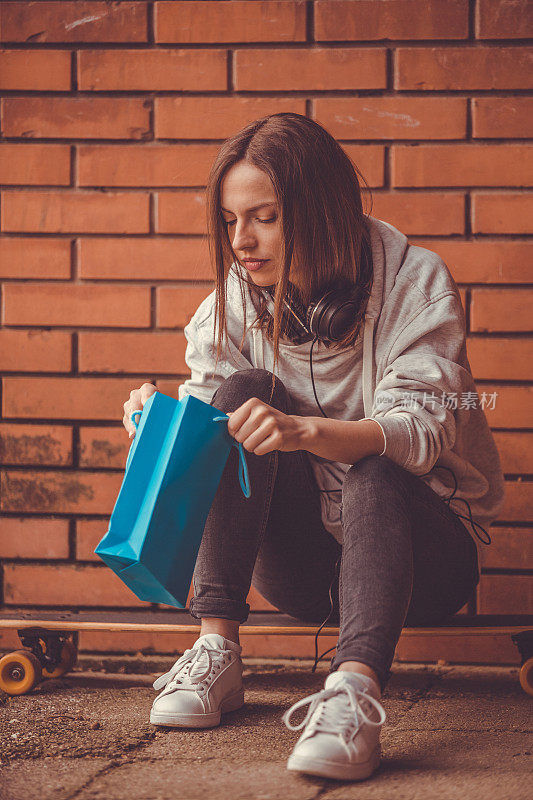 女孩靠砖墙坐在长板上，手里拿着购物袋