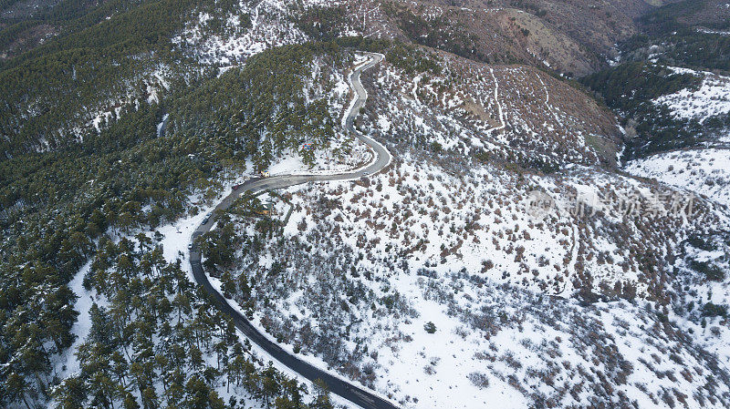 在恶劣天气下，俯视图无人机在弯曲的道路、风山和覆盖着积雪的森林上空飞行。