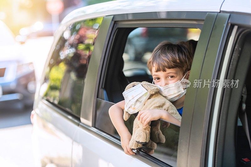 小男孩戴着防护面罩，抱着一只泰迪熊，透过车窗看镜头