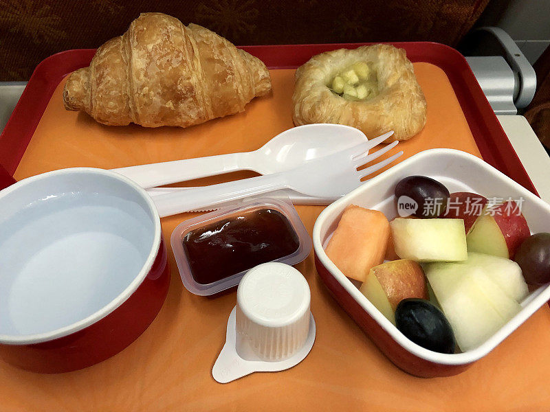 飞机餐盘的特写图片，里面有羊角面包、草莓酱、一碗水果沙拉和一个丹麦苹果，一次性塑料餐具，高架视野
