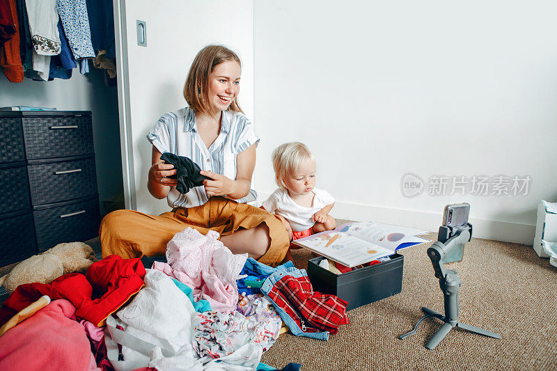 年轻的妈妈在为社交媒体做视频直播，同时在家整理衣服。在智能手机上与朋友和家人进行视频通话。用万向三脚架拍视频和打电话的女人