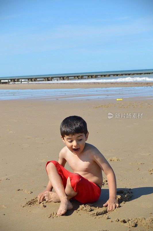 一个穿着红色短裤的小孩张着嘴坐在沙滩上的沙子里