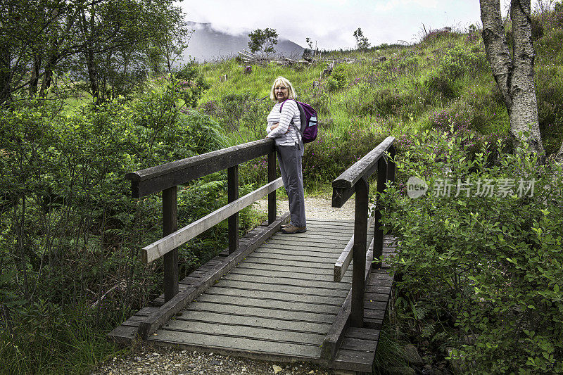 在苏格兰阿德古尔半岛，一位成年女性徒步旅行者在一座木制人行桥上停下来休息，然后回头看向摄像机。