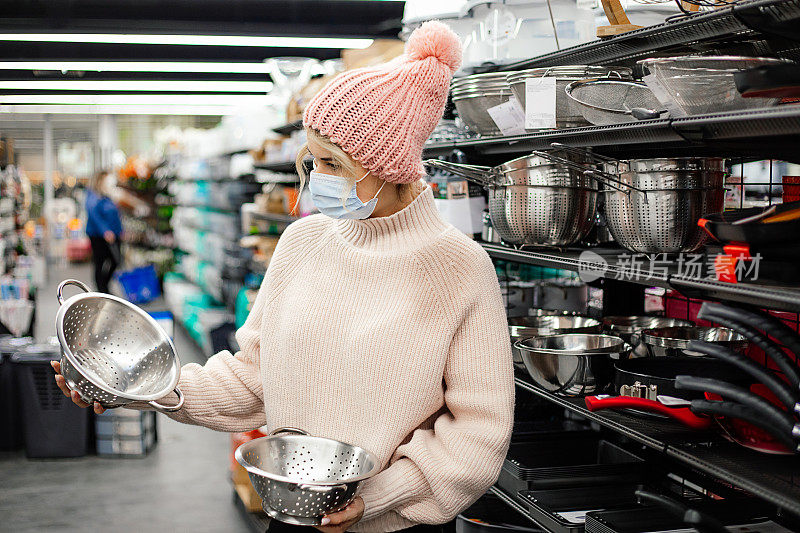 漂亮的顾客在面具，粉红色的针织帽子和毛衣选择两个过滤器在超市的厨房用具部门。购物在大流行。烹饪用具。