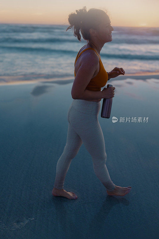 快乐的健身女慢跑者在日落时走在海滩上