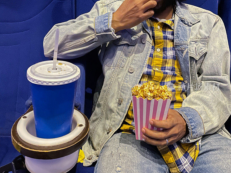 特写图像的人坐在一排电影院座位，拿着硬纸盒焦糖爆米花，一次性饮料杯与饮用吸管在饮料支架，重点在前景
