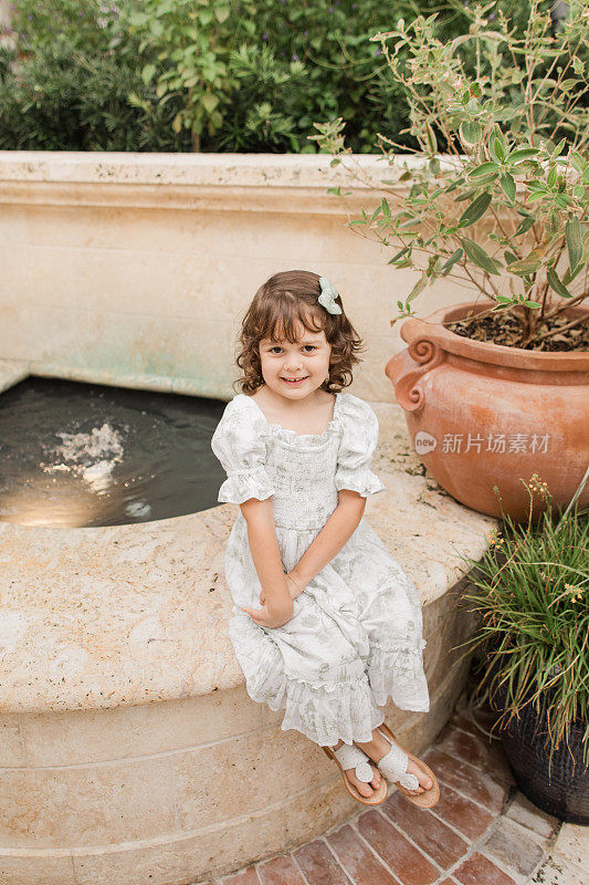 2023年5月，在佛罗里达州棕榈滩的一个托斯卡纳风格的花园中，一个4岁的古巴裔美国女孩，棕色卷发，穿着鼠尾草绿和白色的春季连衣裙，扎着发结，穿着白色的皮凉鞋