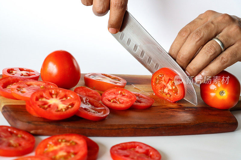 不可识别的人用菜刀在木制砧板上切西红柿(茄)的特写图像，整个，一半和水果片显示薄皮，鲜红的果肉和种子，白色背景，重点在前景