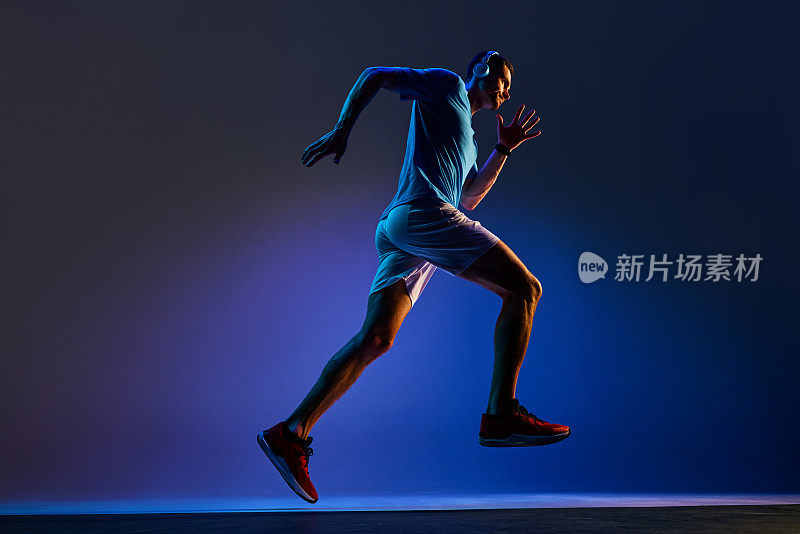 动感的底部视图，肌肉发达的年轻人在运动中，戴着耳机跑步，在霓虹灯的蓝色背景下训练