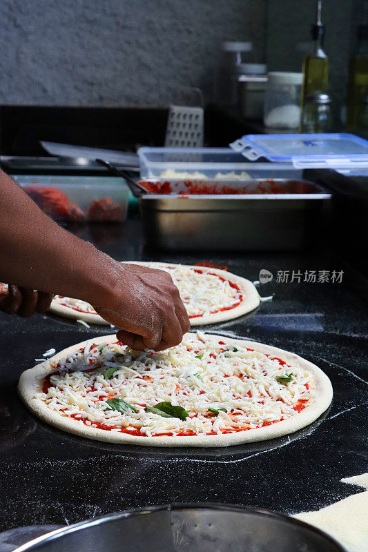 面目全非的Pizzaiolo(披萨厨师)在未煮熟的披萨基础上加入撕裂的罗勒叶，玛格丽塔披萨富含番茄马里纳拉酱，磨碎的马苏里拉奶酪，商业厨房设置，重点放在前景