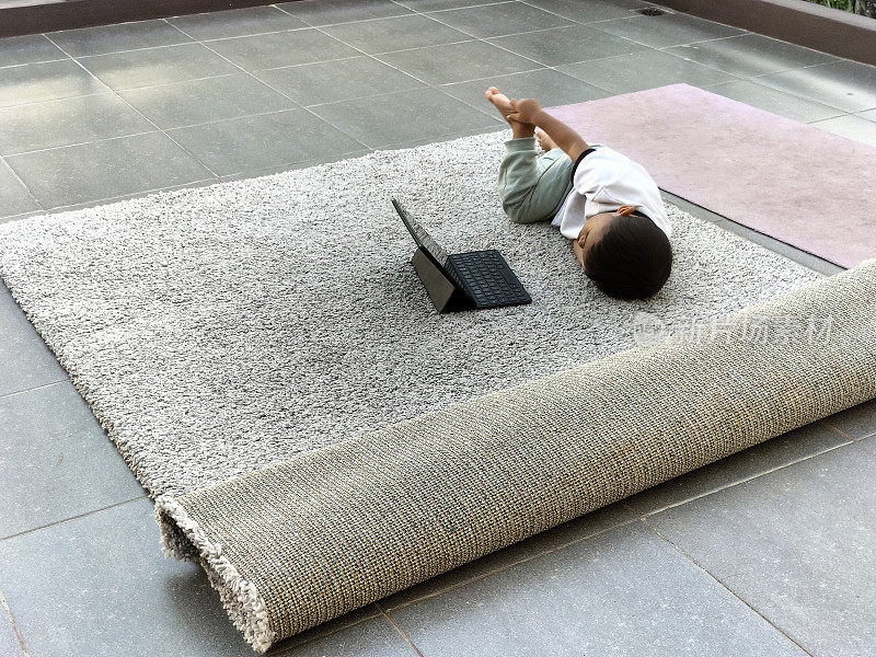 一个年幼的孩子躺在有质感的灰色大地毯上，舒舒服服地使用着笔记本电脑。