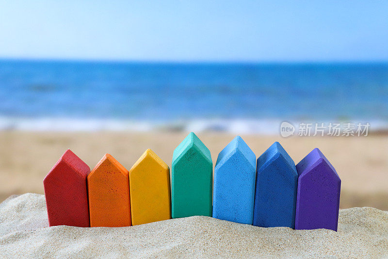 一排彩虹木制模型度假小屋的特写图片，在一堆沙子上，红色，橙色，黄色，绿色，浅蓝和深蓝，紫色，沙滩背景，海浪在退潮时破碎，大海和海岸线，度假租赁和度假屋的概念
