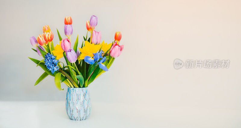 清新的春天色彩柔和的郁金香，水仙花，蝴蝶花在花瓶中站在白色的表面和浅米色的背景。节日鲜花作为礼物。贺卡模型。宽横幅。