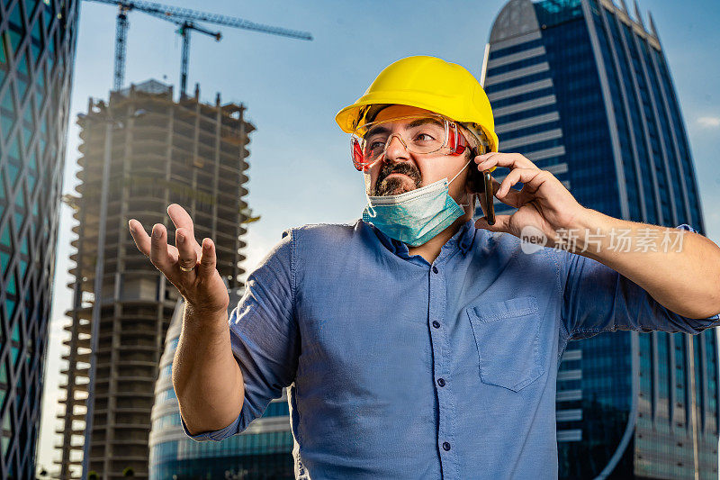 年轻工程师戴着黄色安全帽和N95防护面具在现代商业建筑和建筑工地前使用智能手机
