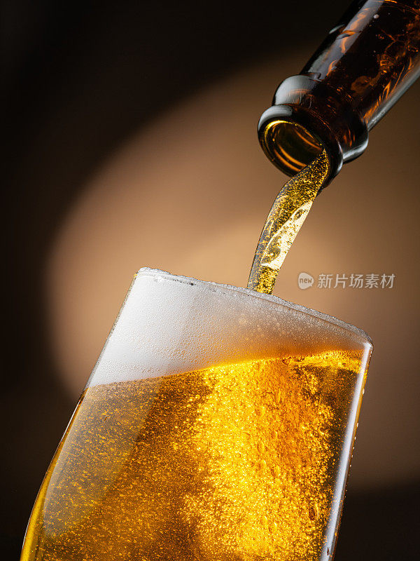 喷出的啤酒从瓶子里倒进啤酒杯里，引起很多的气泡和泡沫。深棕色的背景。