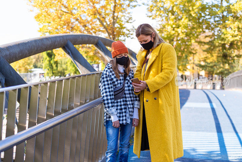 母亲和女儿戴着口罩，在户外行走时使用智能手机