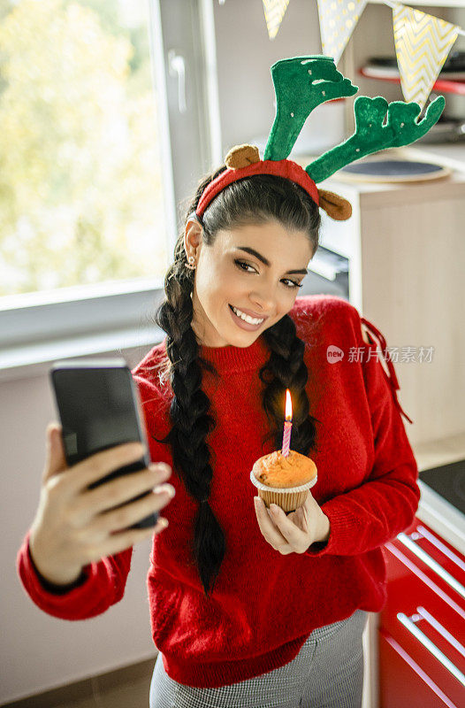 戴着圣诞老人帽的年轻女士正在许愿，拿着带蜡烛的纸杯蛋糕，在厨房里视频通话