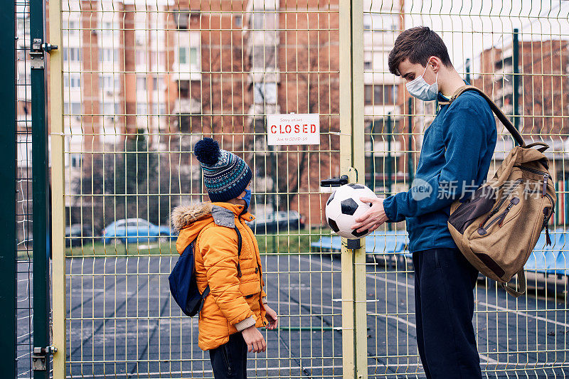 两个男孩在紧闭的操场门前玩球。Covid-19大流行。