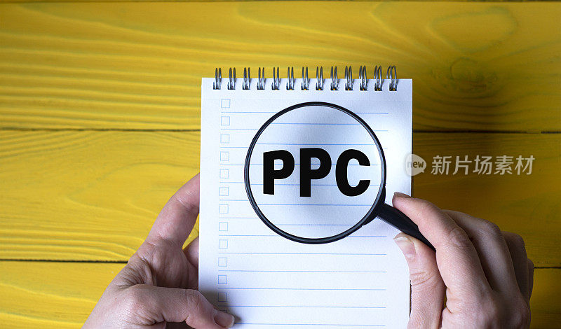 一个女人的手拿着放大镜在一个黄色木制背景上的首字母缩写PPC上。